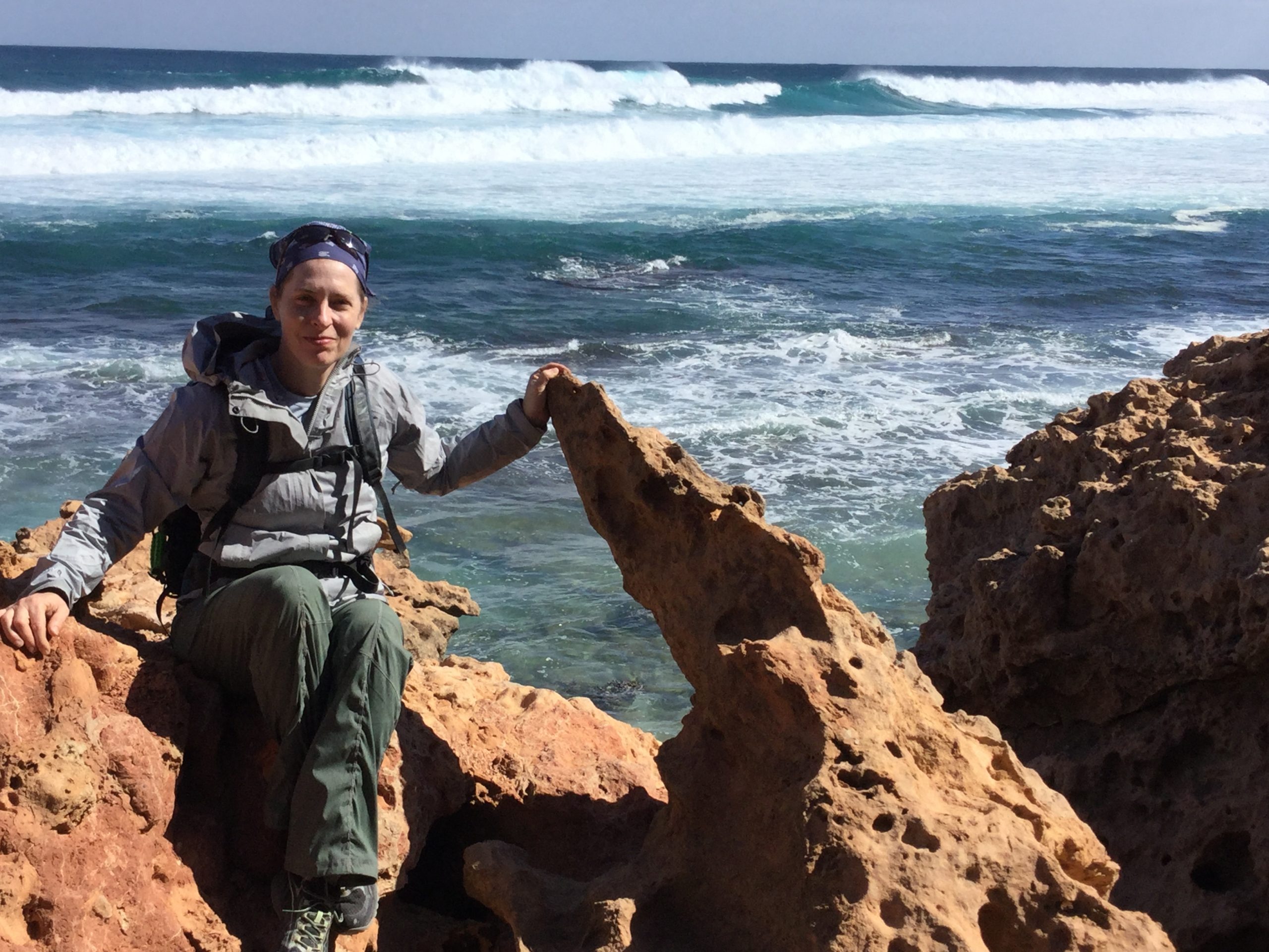Andrea Dutton, sitting on a red rocky outcrop next to the ocean